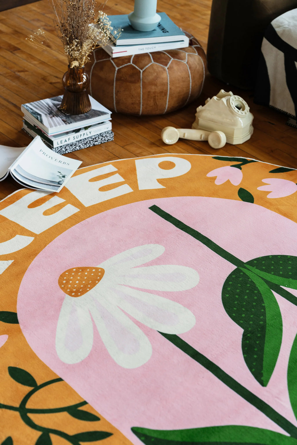 Playful rug with daisy design spread across wood floor in the living room corner with books, pouf, and decor