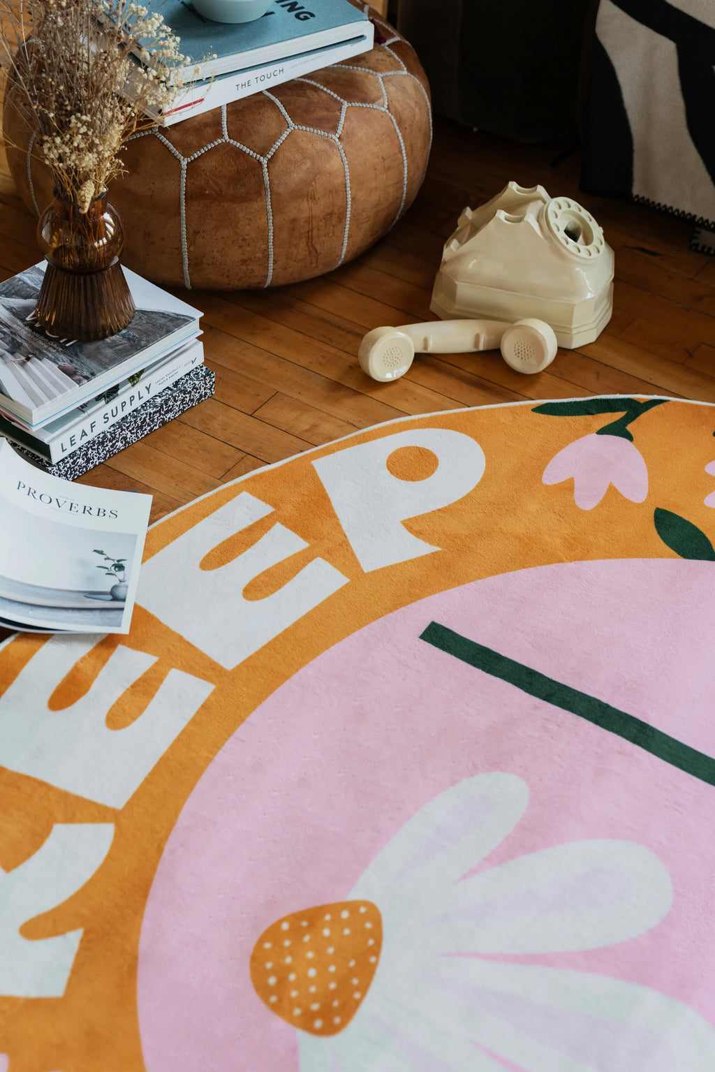 Daisy rug with partial “KEEP” text and floral design on wooden floor beside books, pouf, and vintage phone in living room