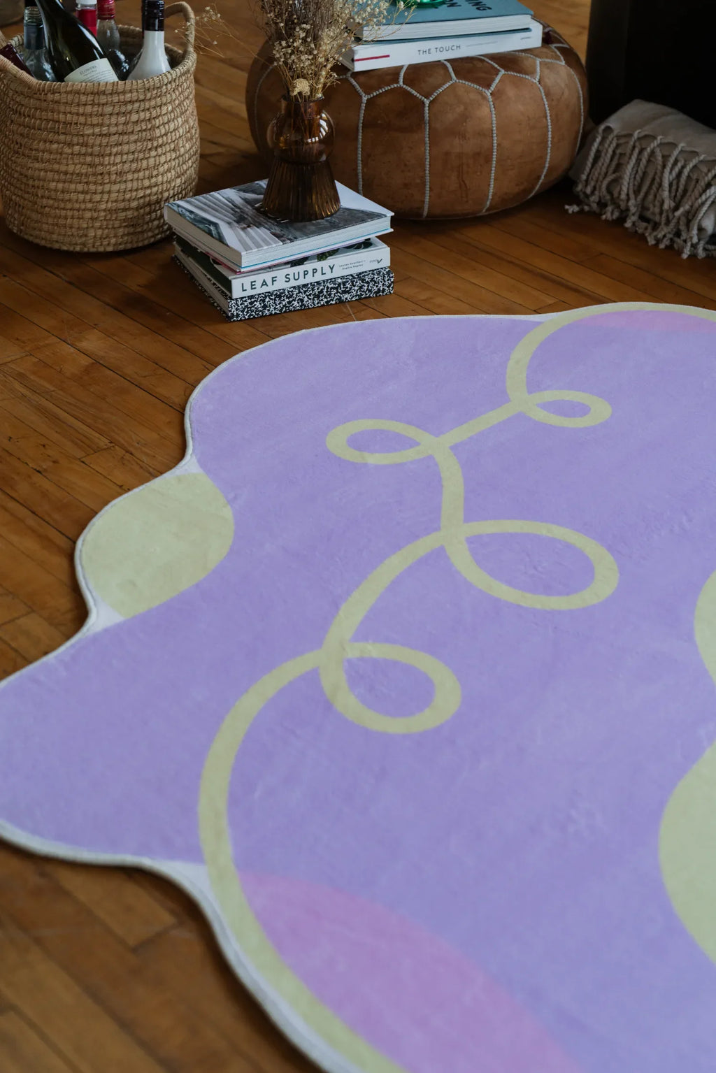 Close-up of lilac rug with playful yellow swirls and scalloped edges, placed on wooden floor beside stacked books and woven pouf.