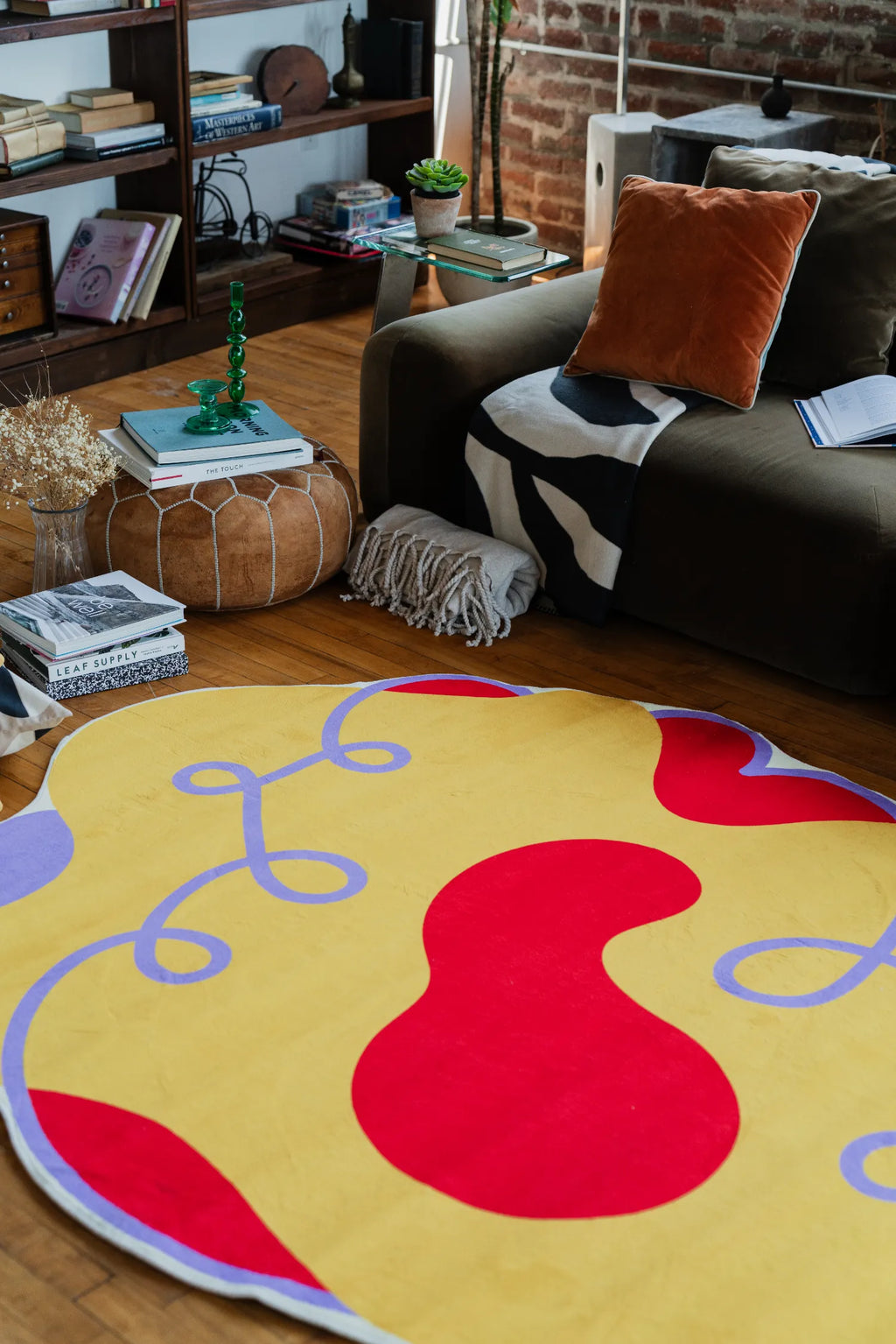 Modern area rug in bold yellow and red hues spread near a sofa with velvet cushions and a stacked leather pouf.