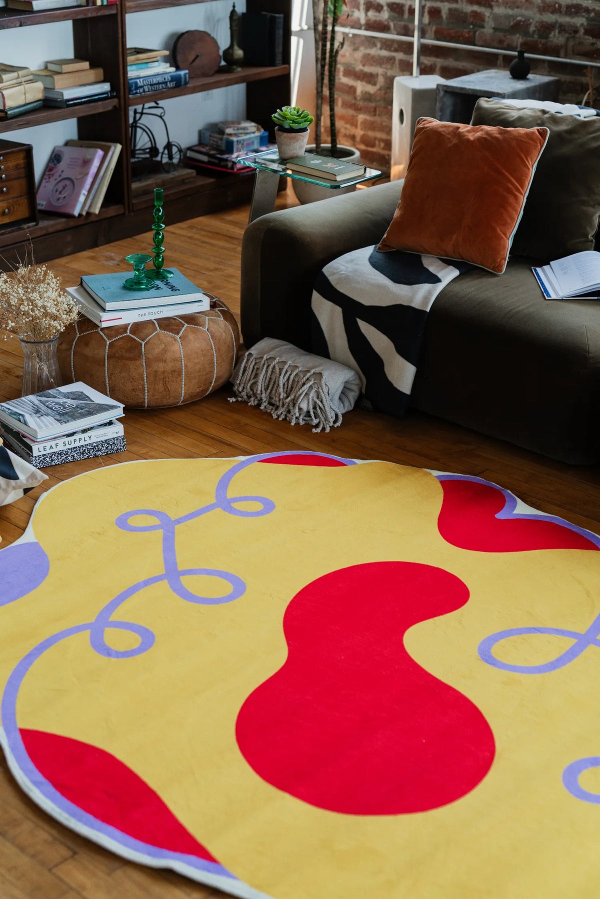 Modern area rug in bold yellow and red hues spread near a sofa with velvet cushions and a stacked leather pouf.