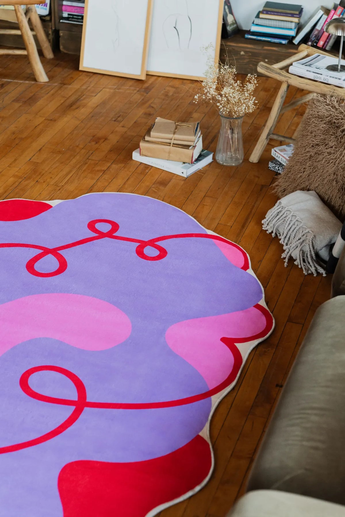 Wavy Abstract Irregular Area Rug by Shayla Goethe in lilac and pink, placed beside a small wooden stool with books and framed art in a cozy corner.