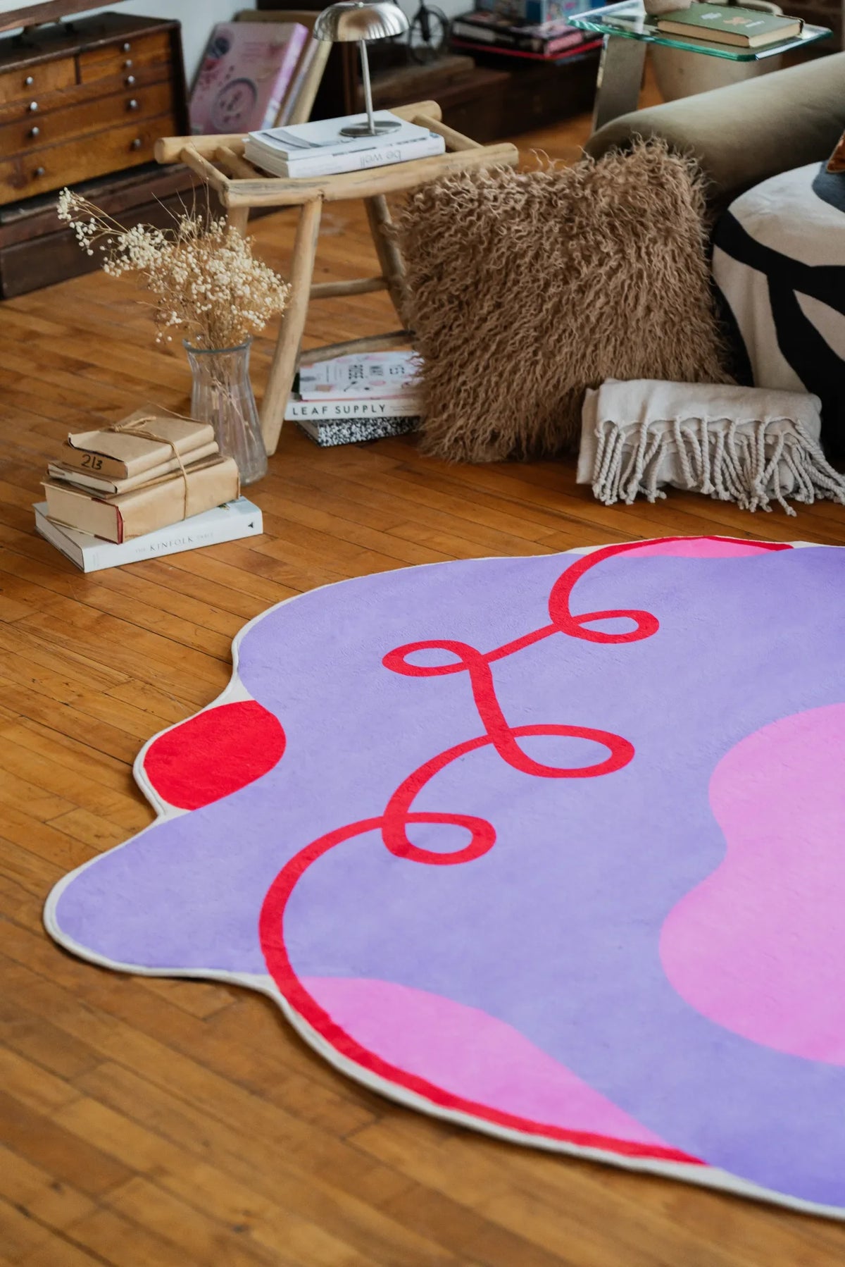Close view of the irregular area rug in lilac and pink, showing its flowing shape and red swirl details next to a sofa and textured cushions.