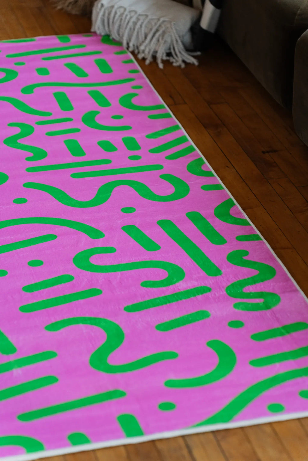 A pink and green retro rug stretched along wood floor beside a seating area with soft textures.