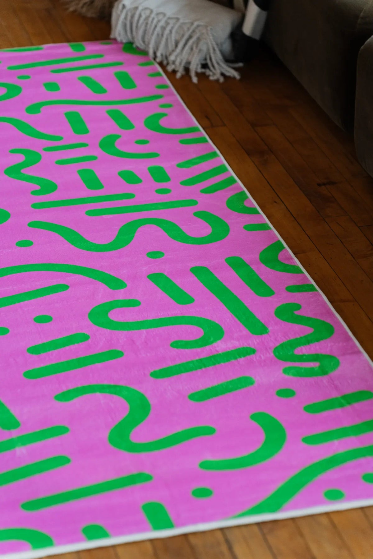 A pink and green retro rug stretched along wood floor beside a seating area with soft textures.