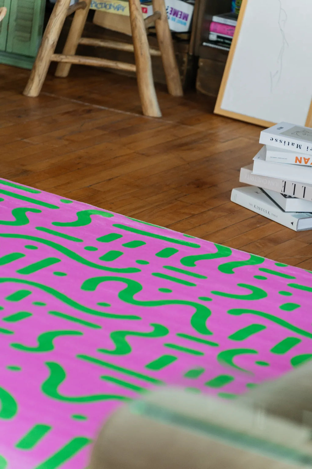Pink and Green Retro Graphic Area Rug by Shayla Goethe placed near stacked books and wooden stool on wood floor.
