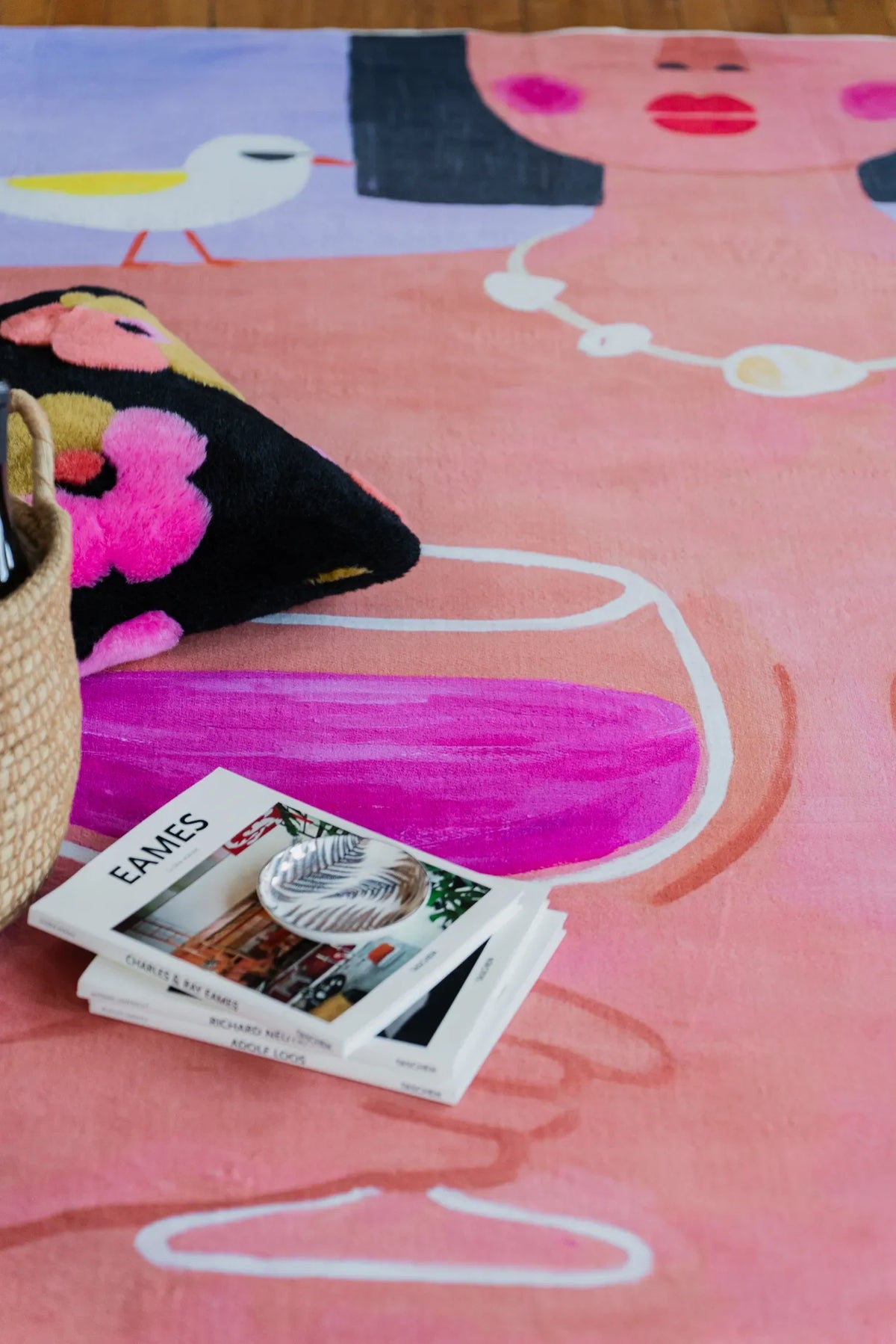 Funky Wine and Woman Art Rug styled with floral cushion and design books resting on wood floor beside basket.