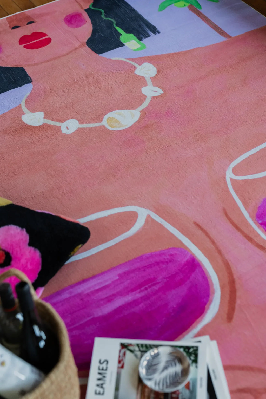 Playful rug showing woman necklace and wine glasses partly covered by basket with bottles and bright patterned pillow.