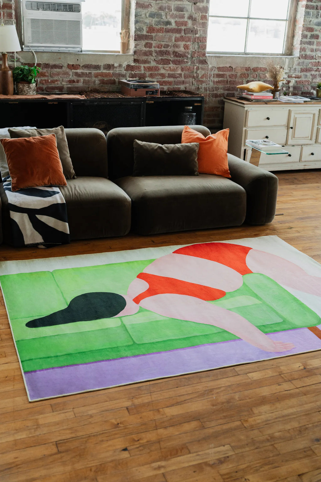 Contemporary loft setting with abstract body art rug in striking color blocks, placed in front of brown sofa accented by orange pillows.