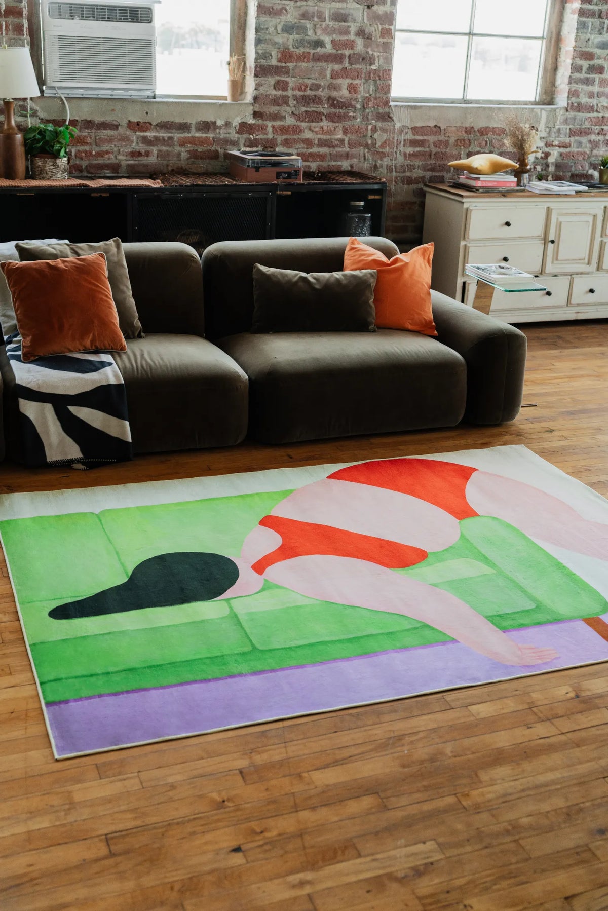 Contemporary loft setting with abstract body art rug in striking color blocks, placed in front of brown sofa accented by orange pillows.