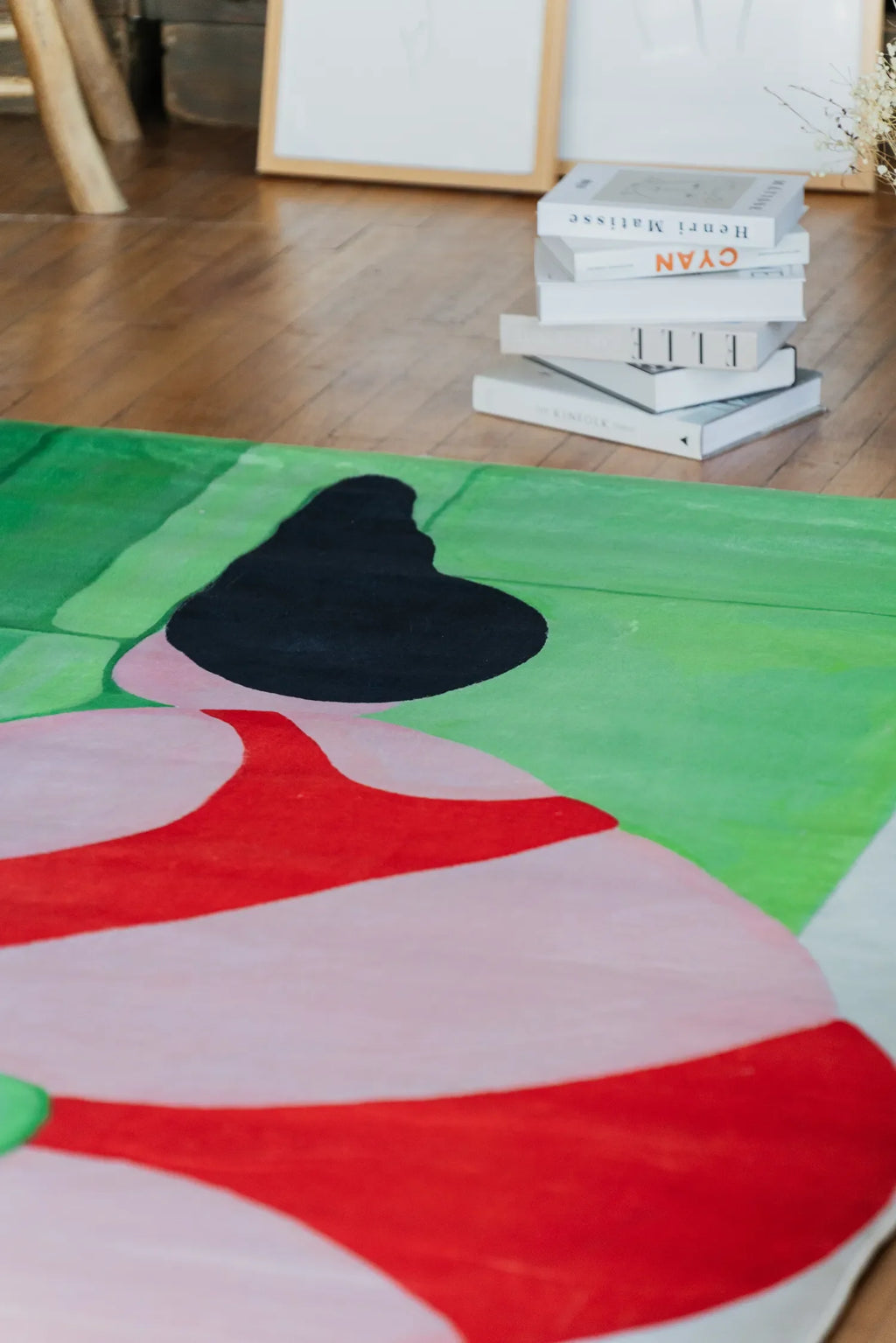 Close-up of modern abstract body art rug with bold red and green tones, books and sketches stacked against the wall in a cozy studio.