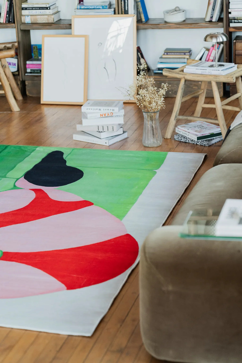 Abstract body art rug displayed near a bookshelf and rustic stool, accented by dried flowers and stacked design books for a warm interior look.