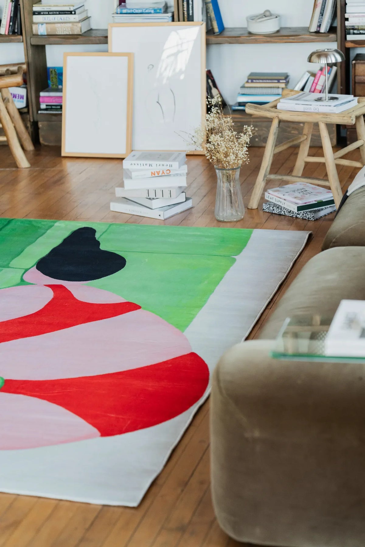 Abstract body art rug displayed near a bookshelf and rustic stool, accented by dried flowers and stacked design books for a warm interior look.