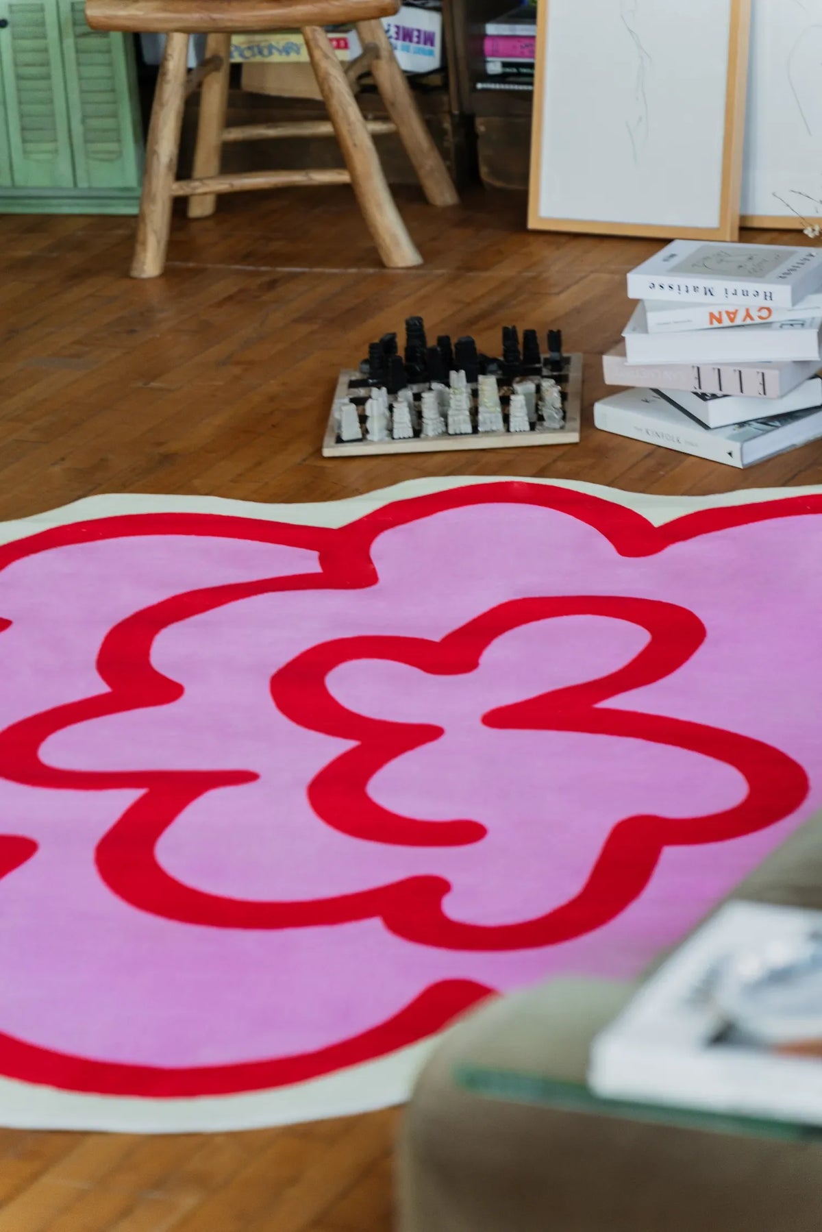 A pink funky flower shaped rug spreads across the living room floor, adding a fun accent near stacked books and a chess set.