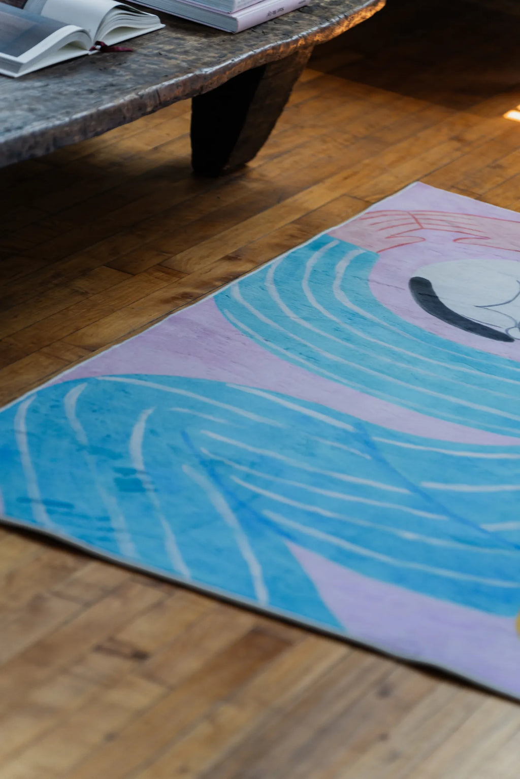 A corner view of the Funky Cat Lover Rug by Nimei in pink, blue, and lilac, styled next to an open book on a bench.