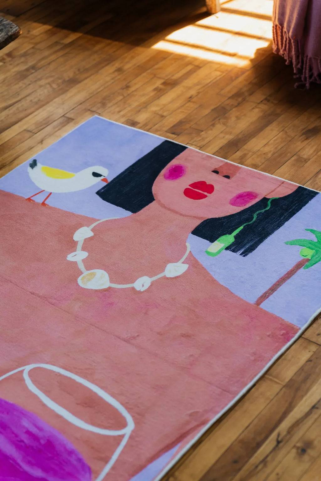 Playful rug with bold woman portrait and pink wine glasses set by soft blanket edge in sunlit corner on wood floor.