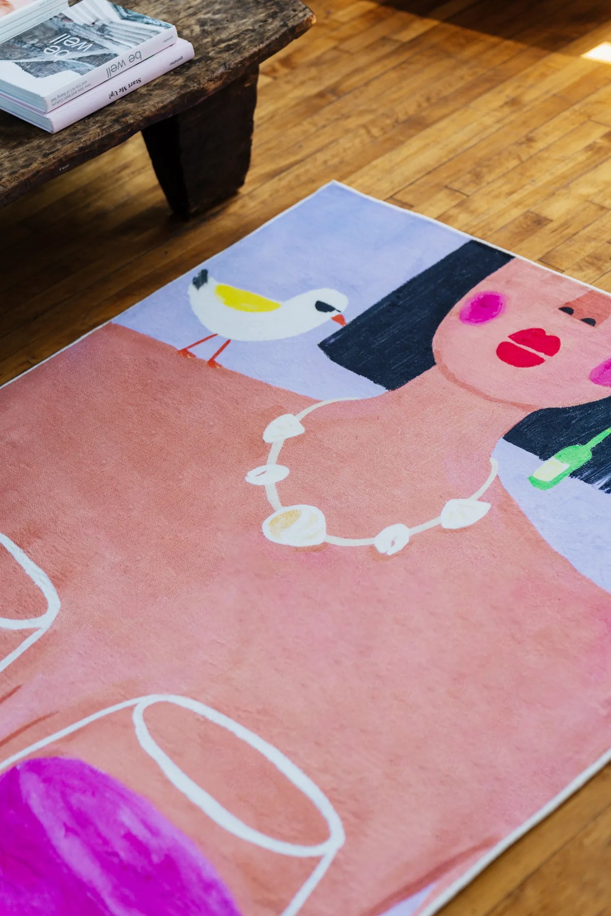 Funky Wine and Woman Art Rug placed near rustic wood bench with design books on warm polished floor.