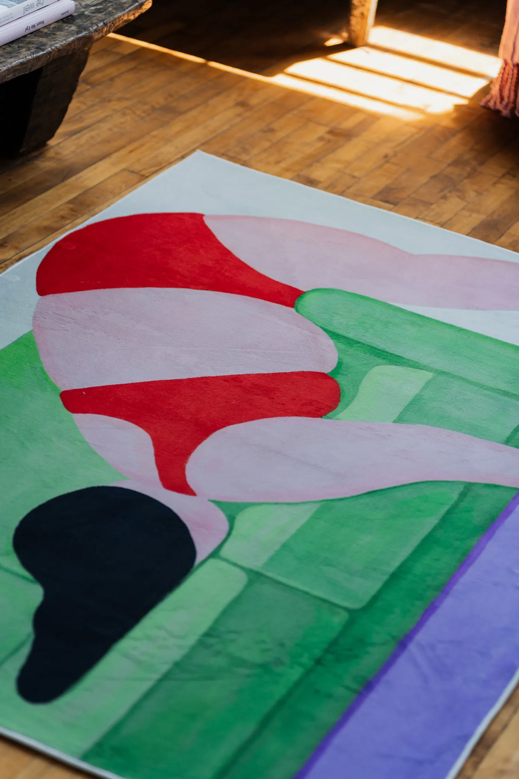 Abstract body art rug in red, green, and lilac tones captured at an angle by a sunlit corner of the loft.