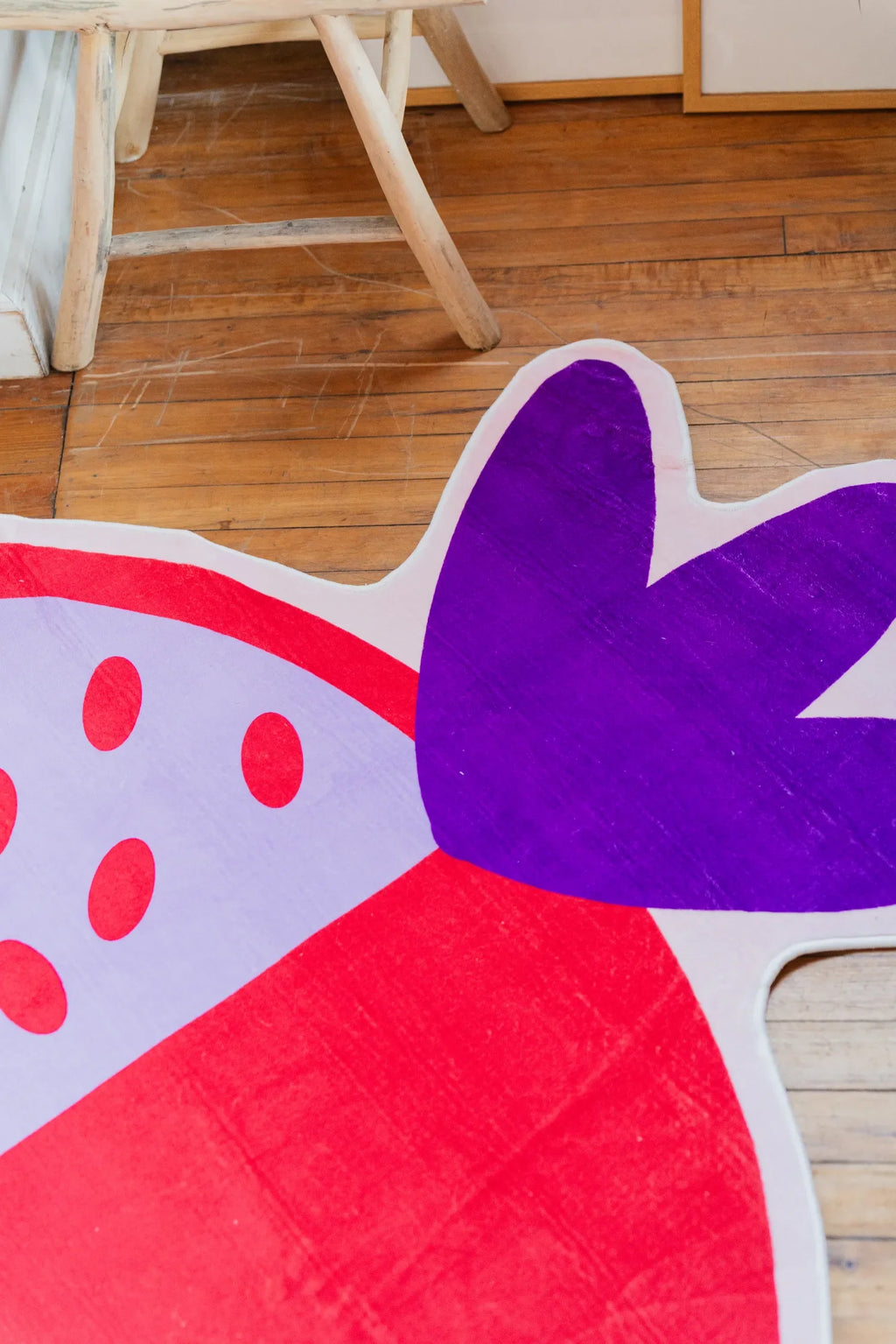 Close-up of pomegranate-shaped rug in vivid red and purple tones, showing playful irregular edges and dotted seed pattern on wooden floor.