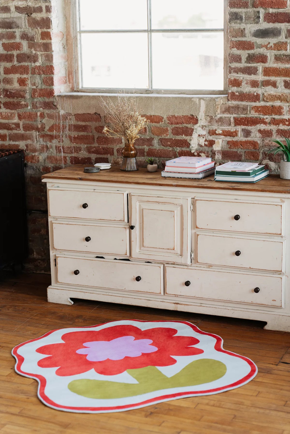 Flower Rug by Anne Julie in bold orange placed near vintage white dresser against exposed brick wall.