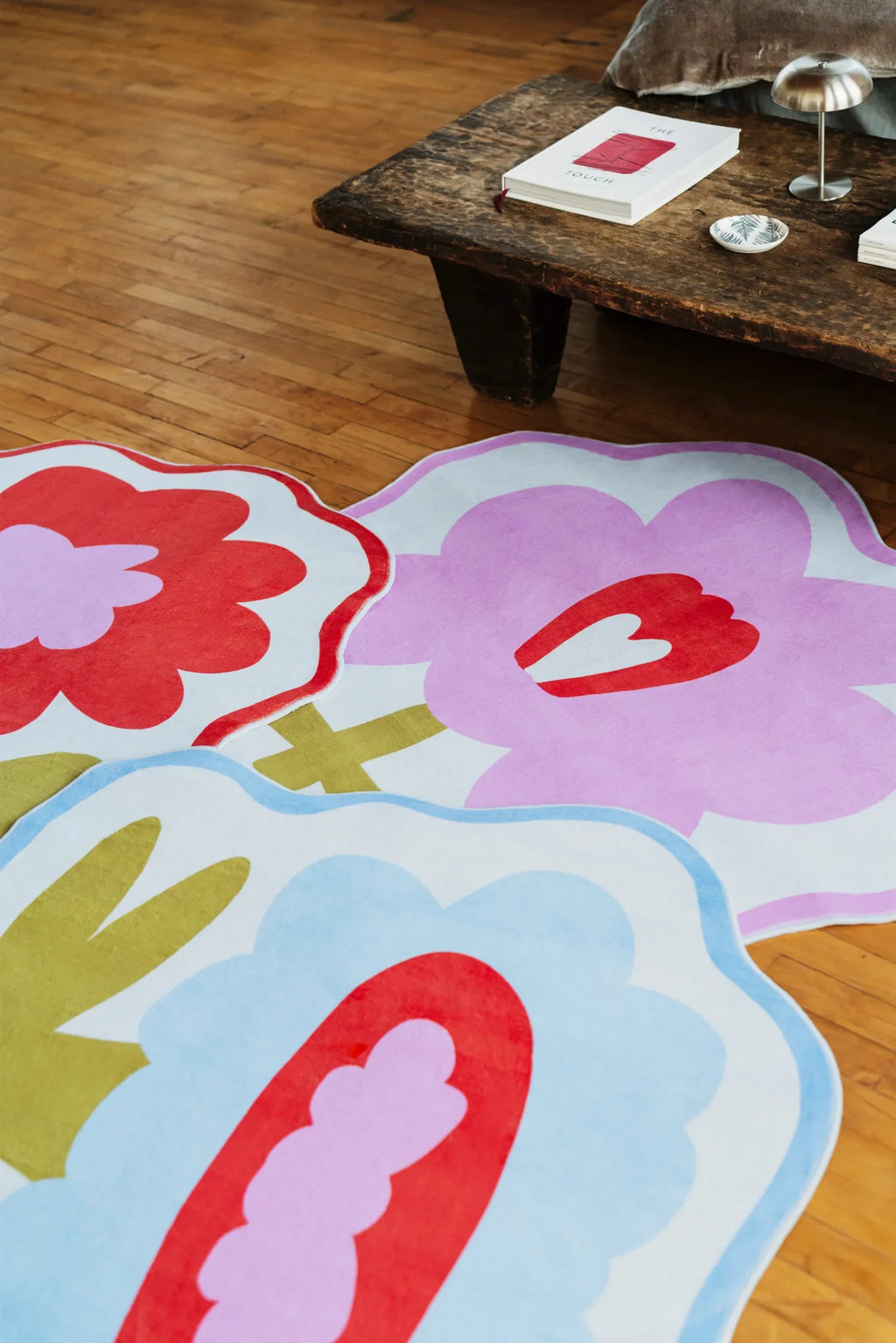 Overhead view of multiple flower-shaped rugs in pink, blue, and orange tones arranged by a wooden bench.