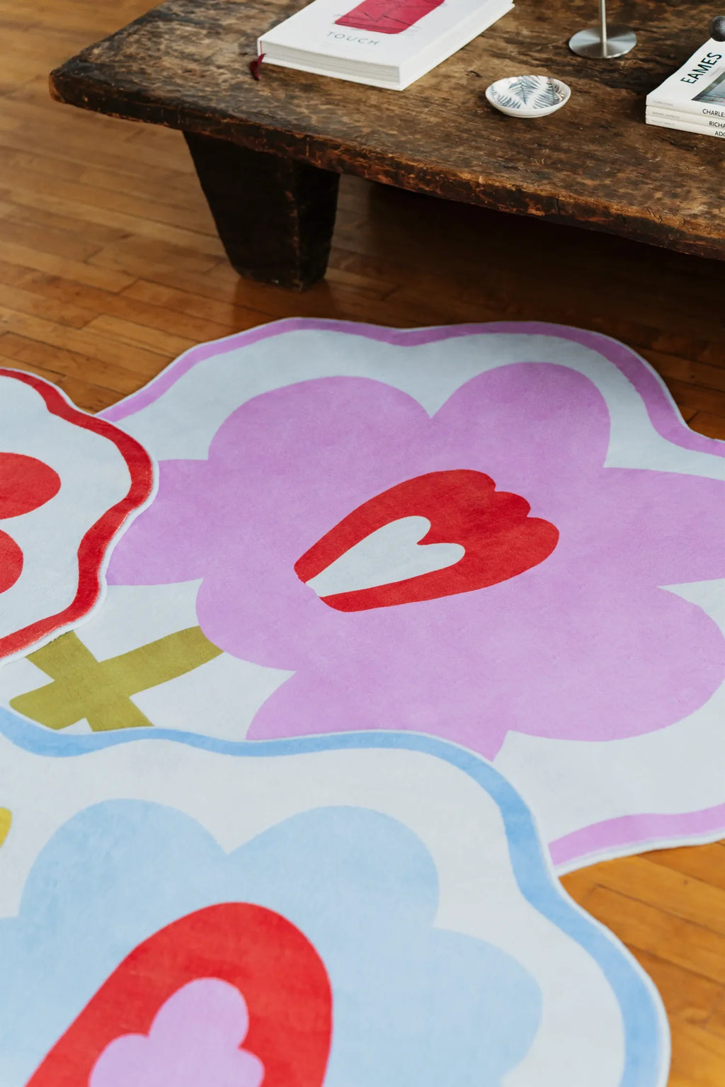 Pink flower rug in focus, layered with blue and orange floral rugs on hardwood floor beside a rustic bench.