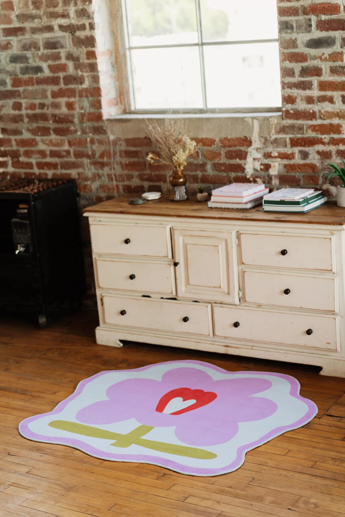 Flower Rug in pink version, placed near a white vintage dresser against exposed brick wall, lit by natural daylight from window.