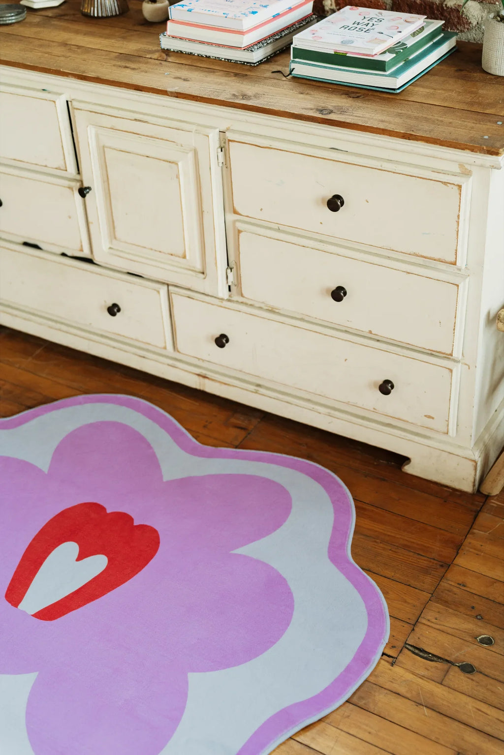Partial view of Flower Rug by Anne Julie in pink version, lying under a distressed white cabinet, captured from a side angle with cozy details.