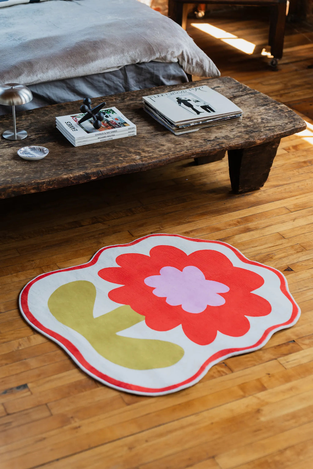 Close-up of Flower Rug by Anne Julie in orange version, styled beside a rustic wooden bench with books and lamp, on warm wooden floor.