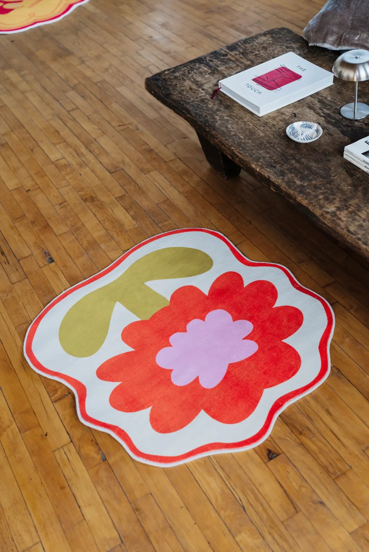 A orange flower-shaped rug with white wavy edges placed on a wooden floor, next to a rustic wooden bench with books and a lamp.