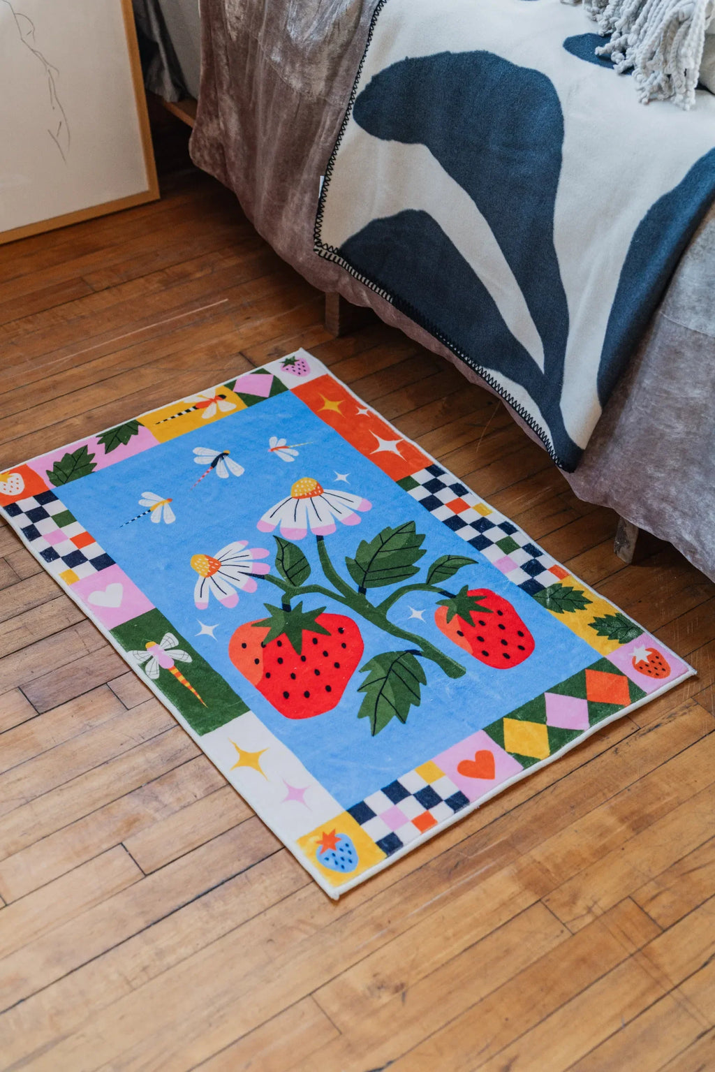 Strawberry Patch Magic Rug styled beside bed on wood floor with cozy blanket draped adding playful bedroom charm