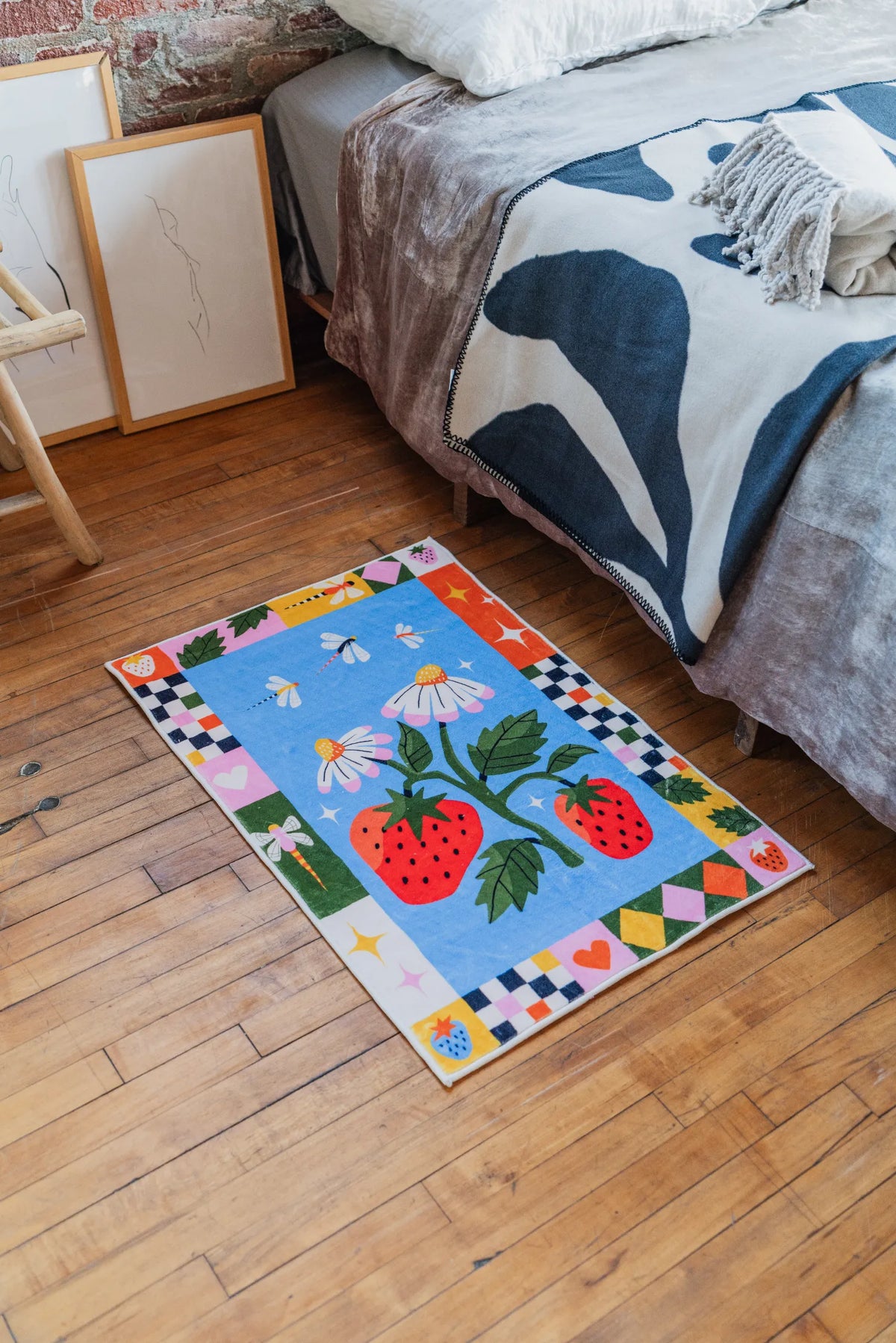 Colorful strawberry rug placed near bed with bold patterned throw and framed art leaning against brick wall creating cozy vibe