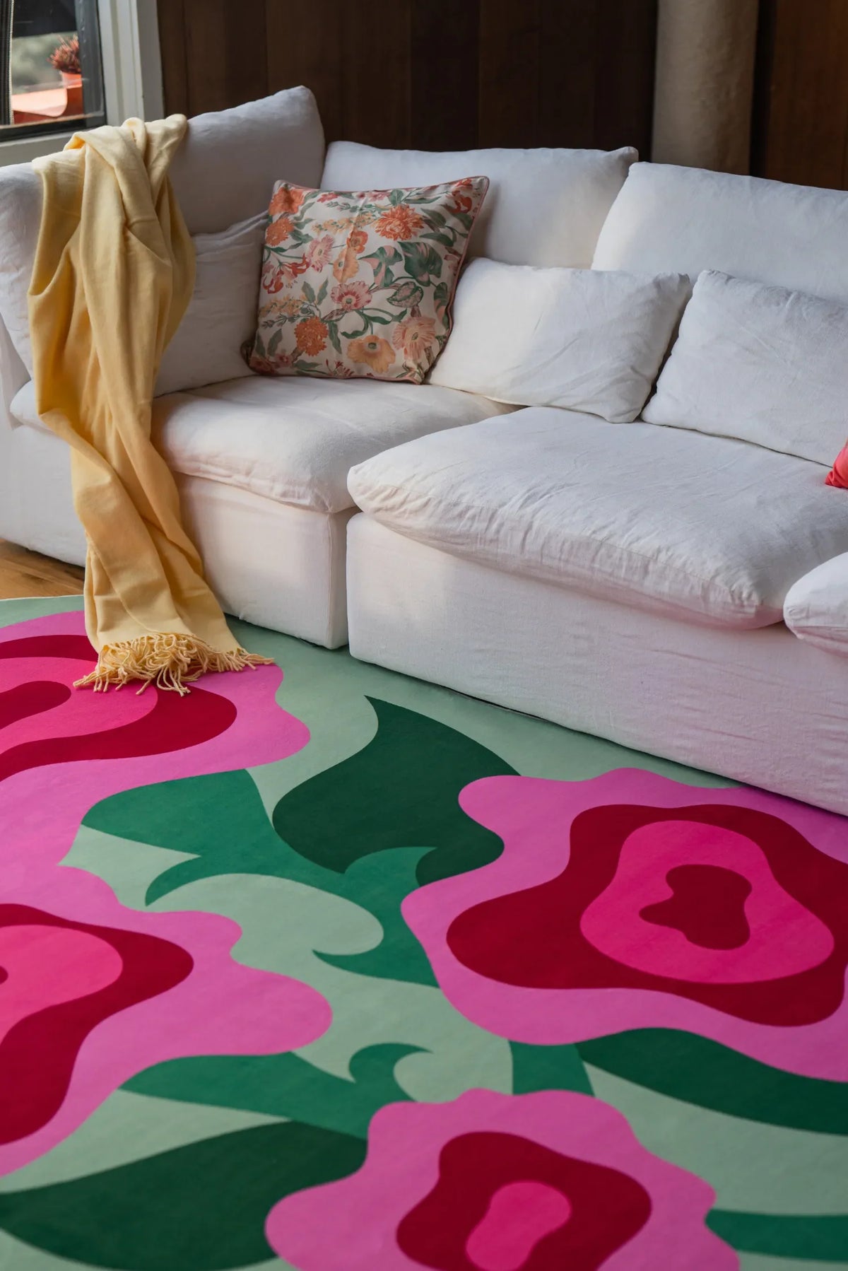 Pink rose rug laid in front of a white sectional sofa with floral pillow and throw, on light wood floor.