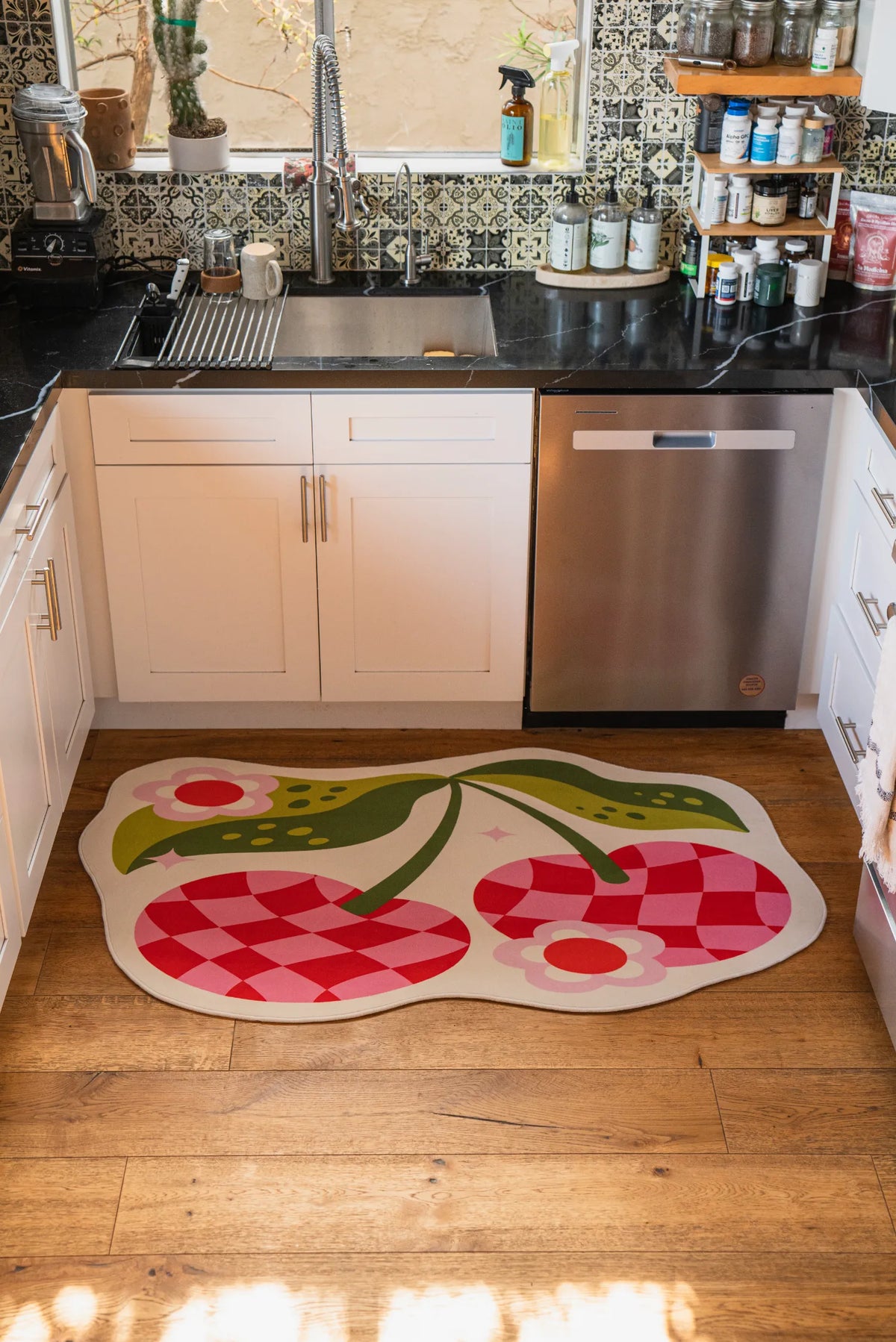Checkered cherry rug set in front of a kitchen sink on natural wood floor between white cabinets.