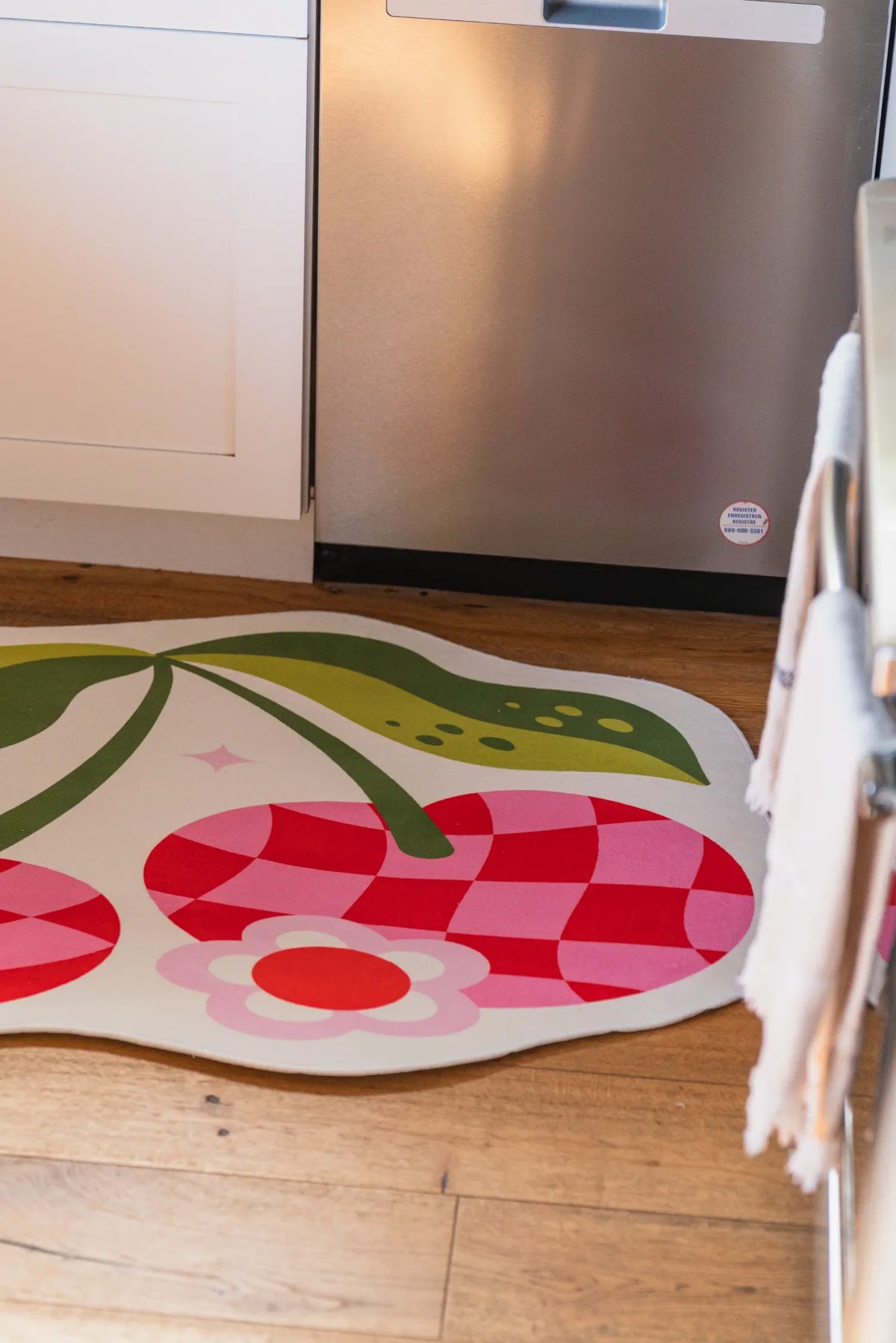 Close view of the pink and red checkered cherry rug beside a stainless dishwasher on wood floor.