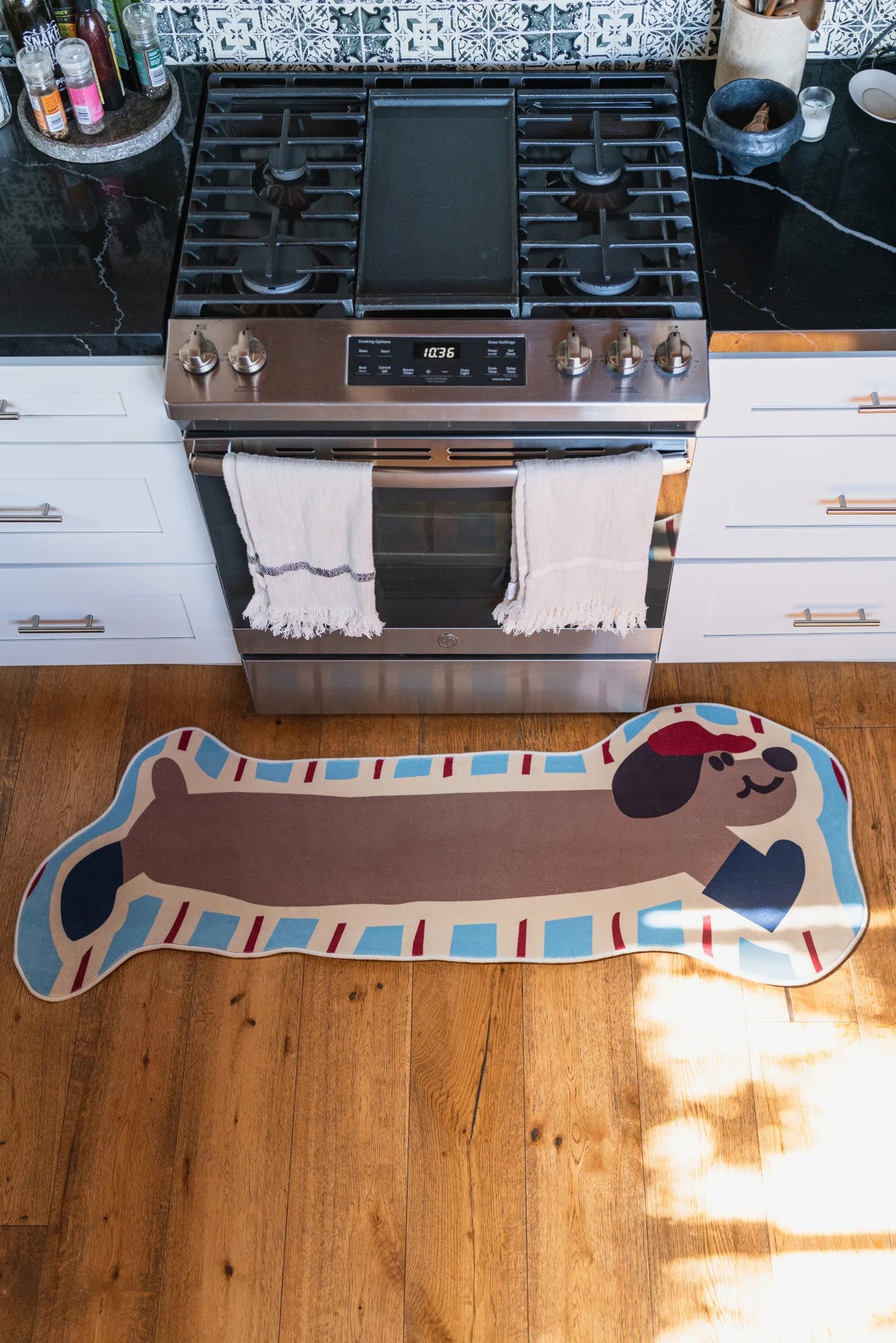 Top-down view of the brown dog rug centered on wood flooring in a kitchen, surrounded by white cabinetry and stove.