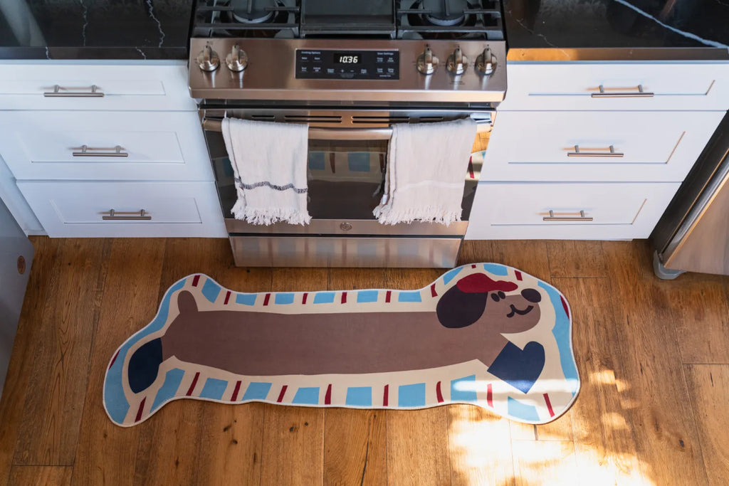 Dog-shaped rug with brown body and blue details placed in front of a kitchen stove on rustic wood floor.