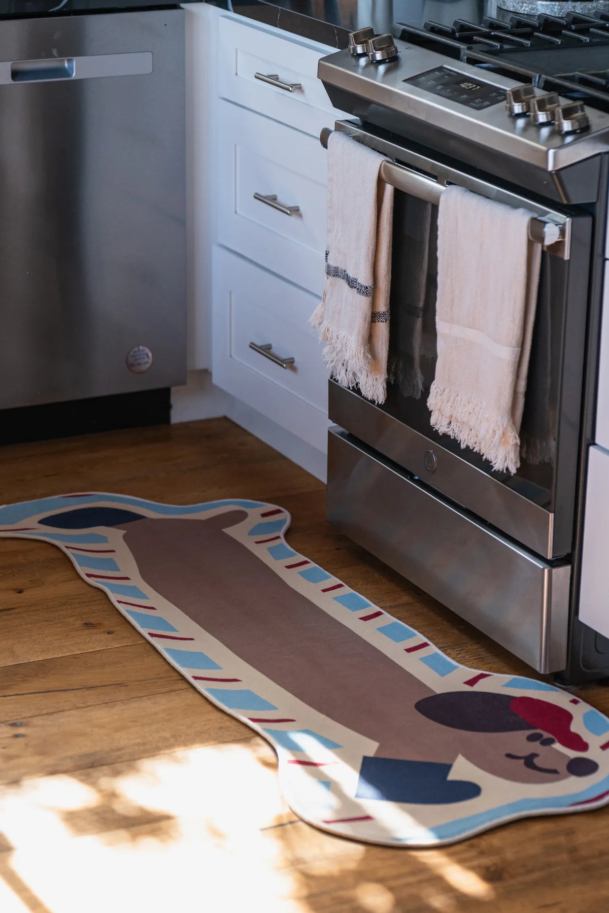 Warmly lit kitchen scene with the brown dog-shaped runner rug laid out on a wooden floor between stove and dishwasher.