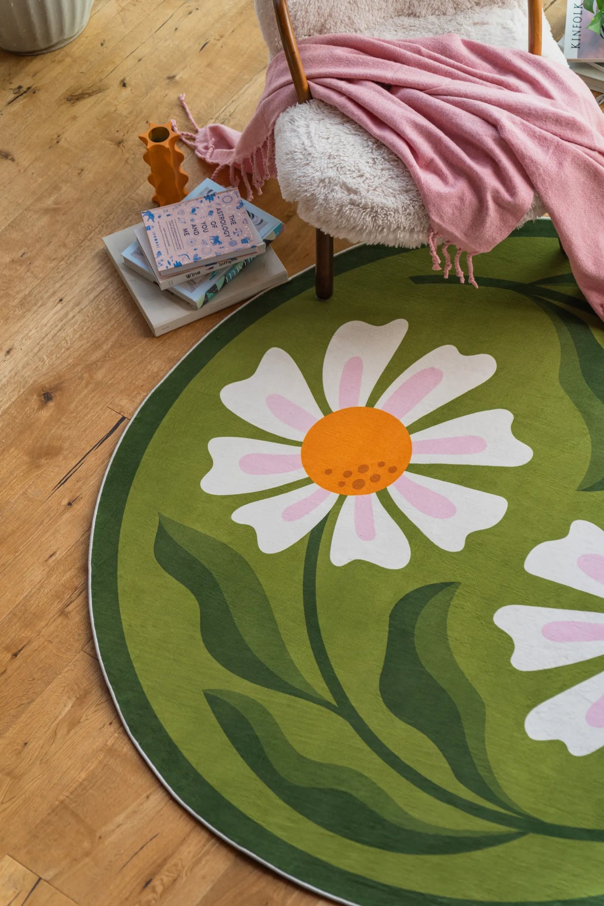 Detailed view of one flower on the green daisy rug, beside stacked books and a pink blanket in the living room.