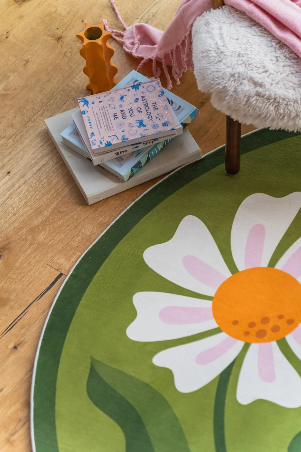 A close-up of the Dual Daisy Round Rug placed under a cozy reading chair with books and a pink throw.