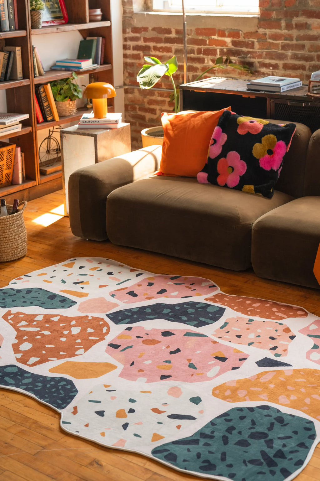 Vibrant terrazzo-style rug in pink, mustard, and teal tones in front of a sofa with colorful pillows and brick wall
