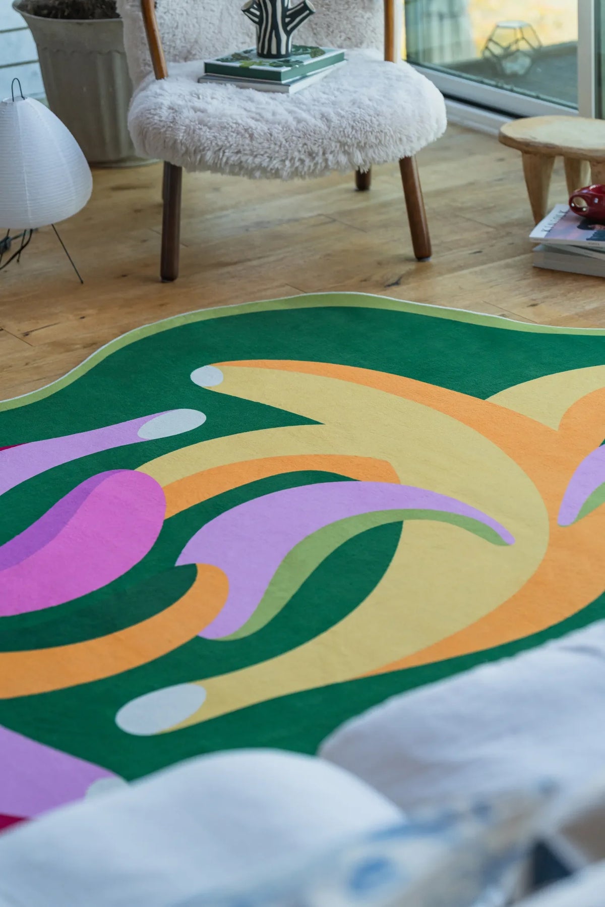 Close-up of funky bananas rug with pink and yellow curves, beside a white sofa and wooden stool by the window.