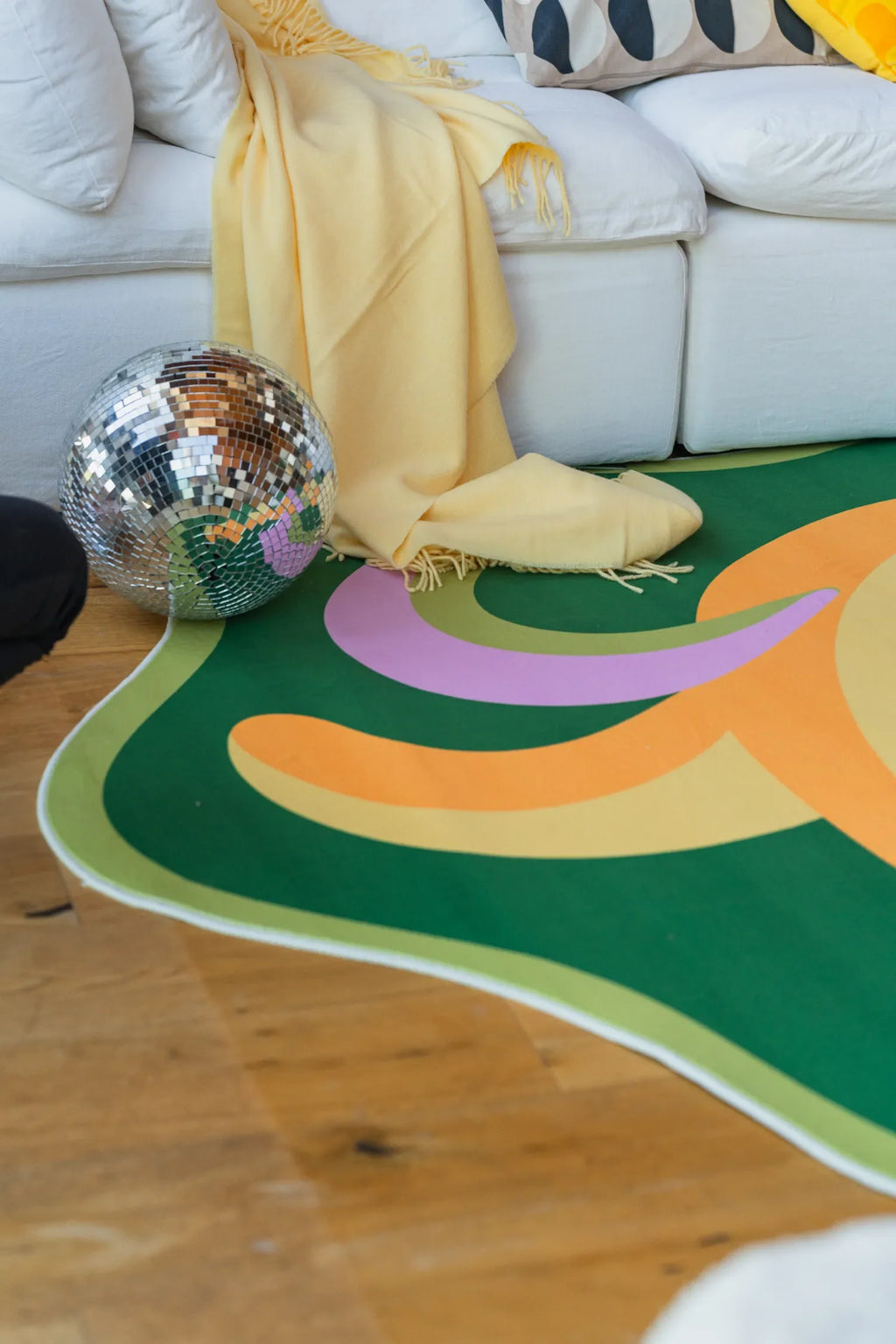 Disco ball and yellow throw placed beside the vibrant funky rug with banana motifs in front of a white couch.