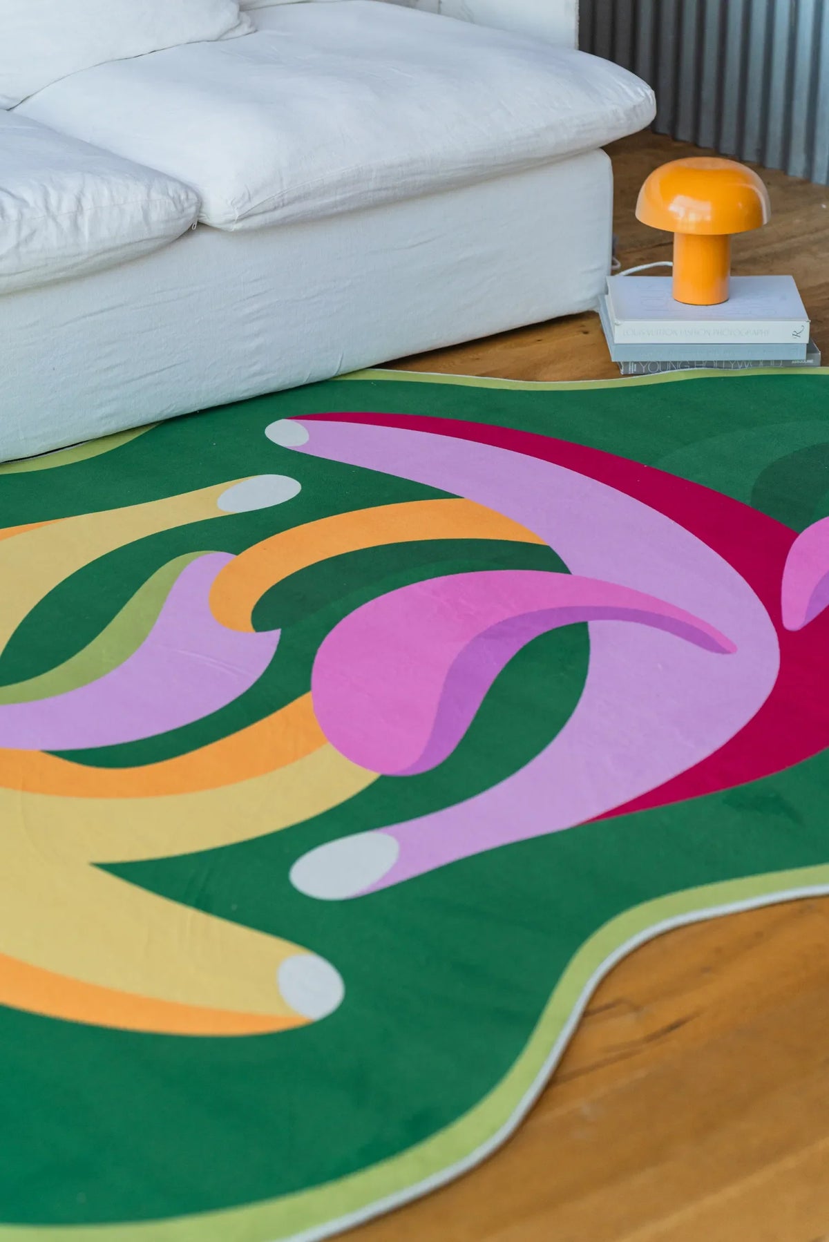 Close-up of the colorful dancing banana rug featuring pink, green, and yellow curves next to a white couch and orange lamp.