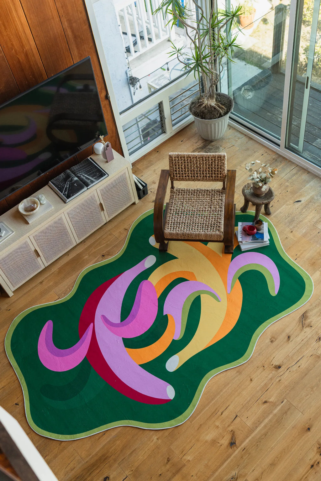 Top-down view of the funky bananas rug in a sunlit corner with a woven armchair, side table, and tall potted plant by the window.
