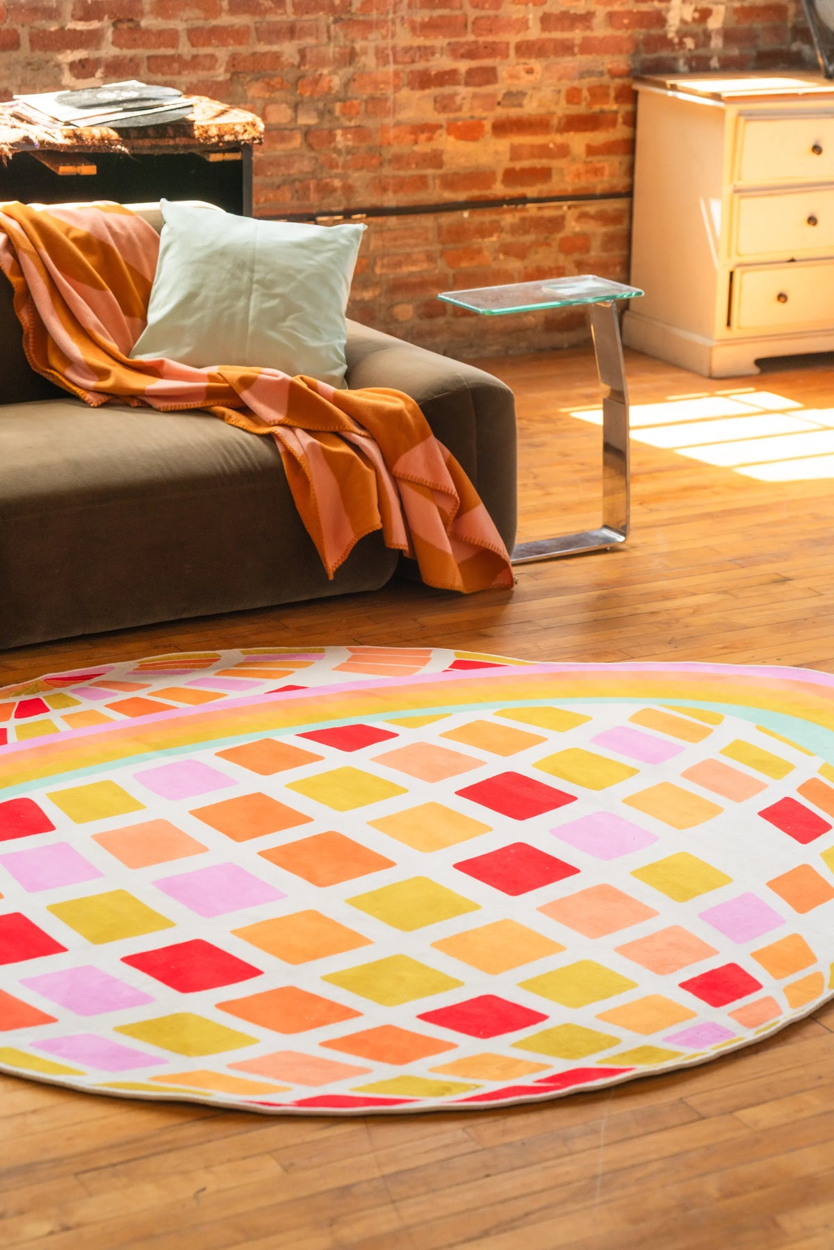 Orange planet checkered rug placed in front of a low sofa with throw, set against warm wood floor and brick wall backdrop