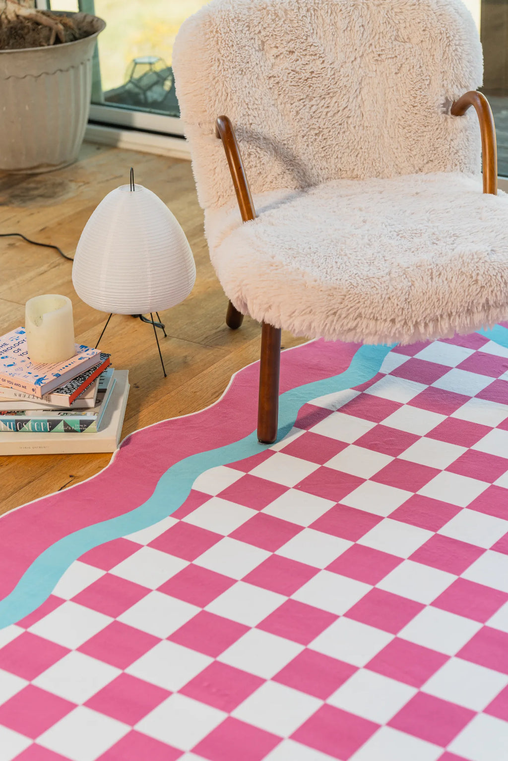 The checkered rug styled under a fuzzy accent chair, paired with stacked books and a soft lighting setup near the window.