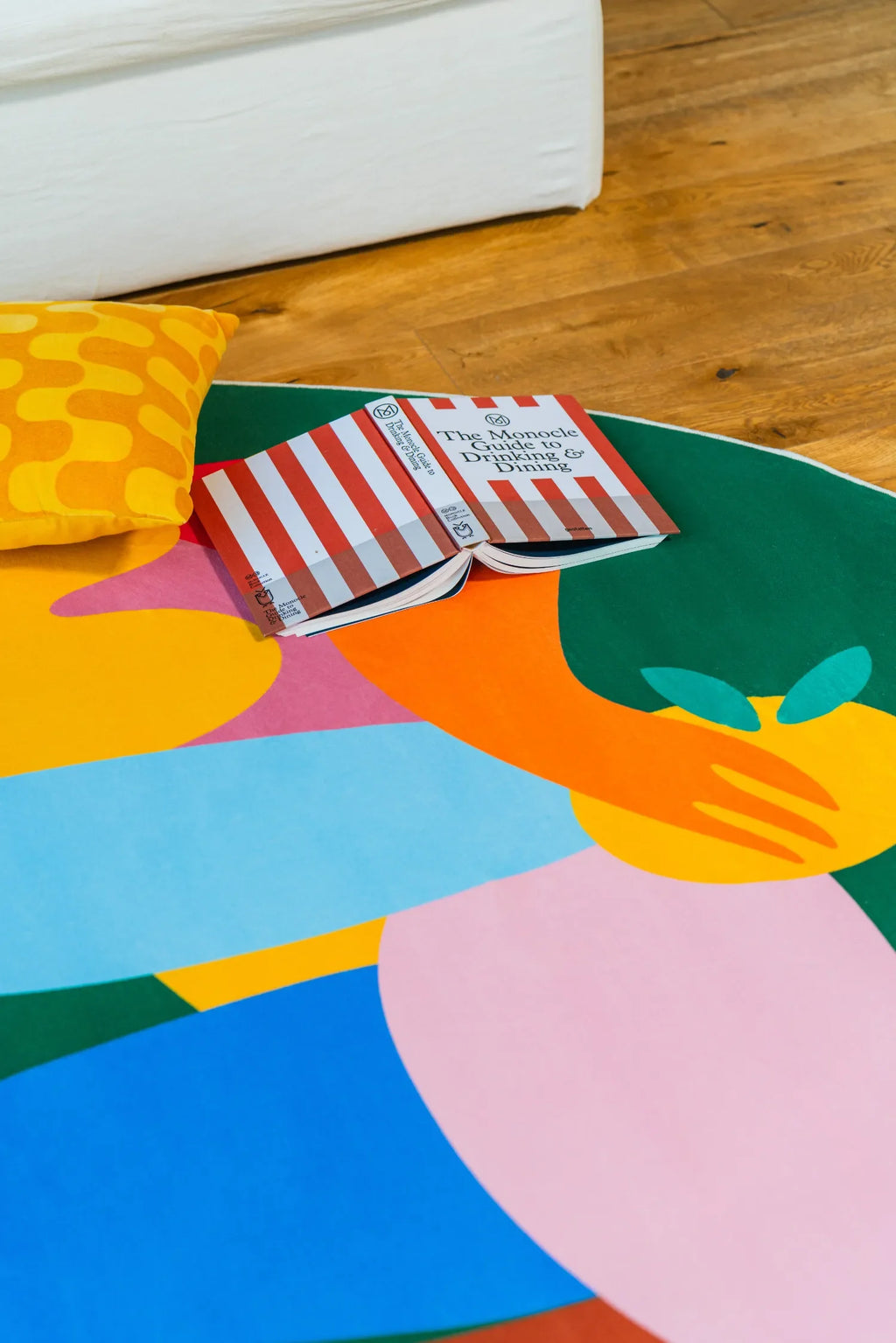 Close-up of green oval rug with colorful fruit-inspired figures beside a cushion and open book on the floor.