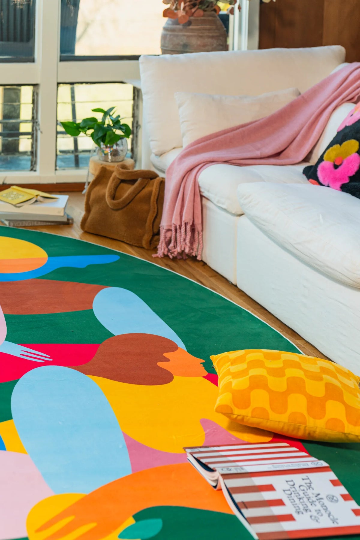 Green oval rug with abstract figures laid out in front of a white couch in a cozy, plant-filled sitting area.