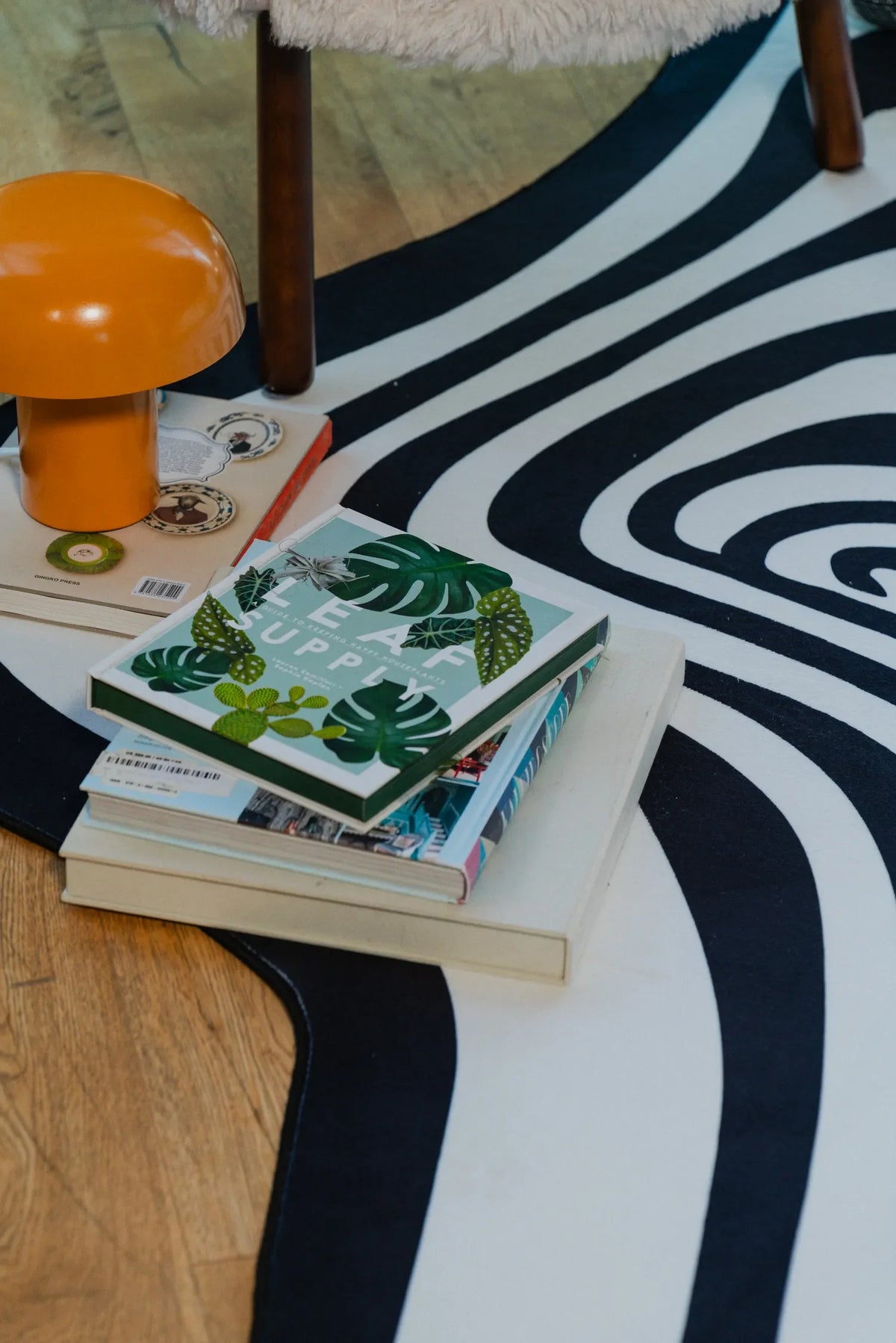 Black and white vertigo rug styled with books and a retro orange mushroom lamp beside a fluffy chair in a cozy reading nook.